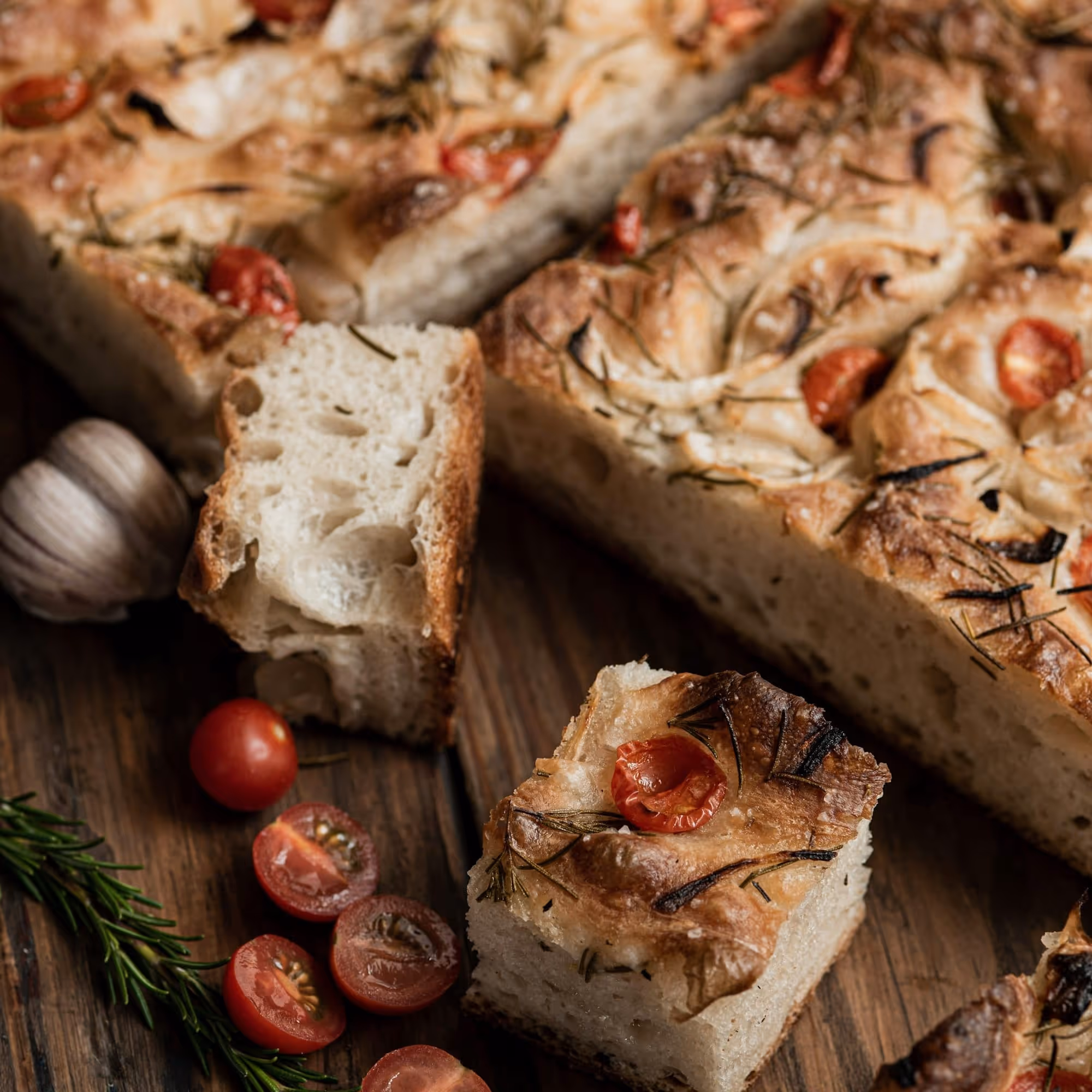 Focaccia barese cut into squares on a dark board with cherry tomatoes, rosemary, and a head of garlic