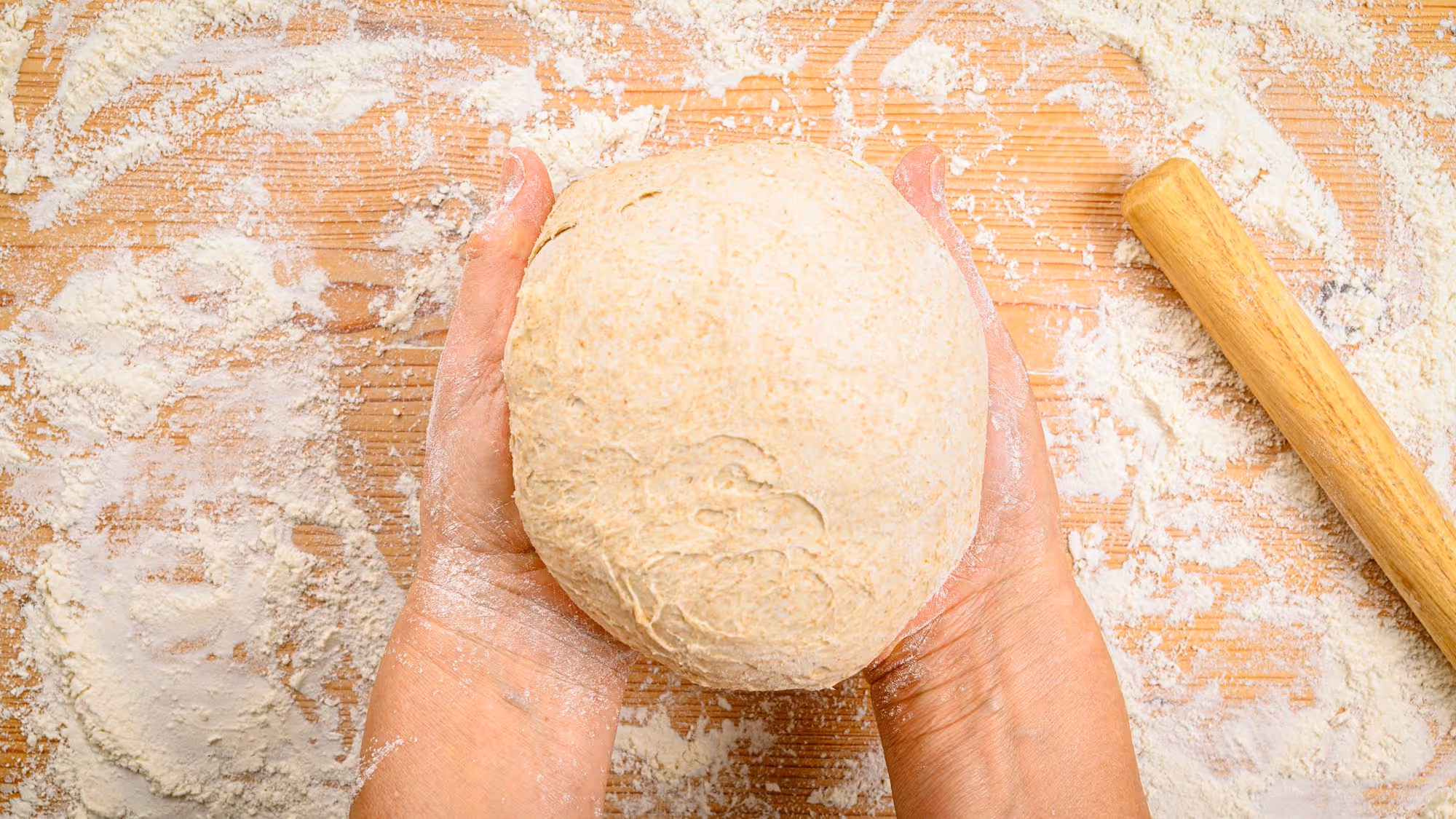 Two hands cradling a rested ball of semolina dough over a floured wooden bench