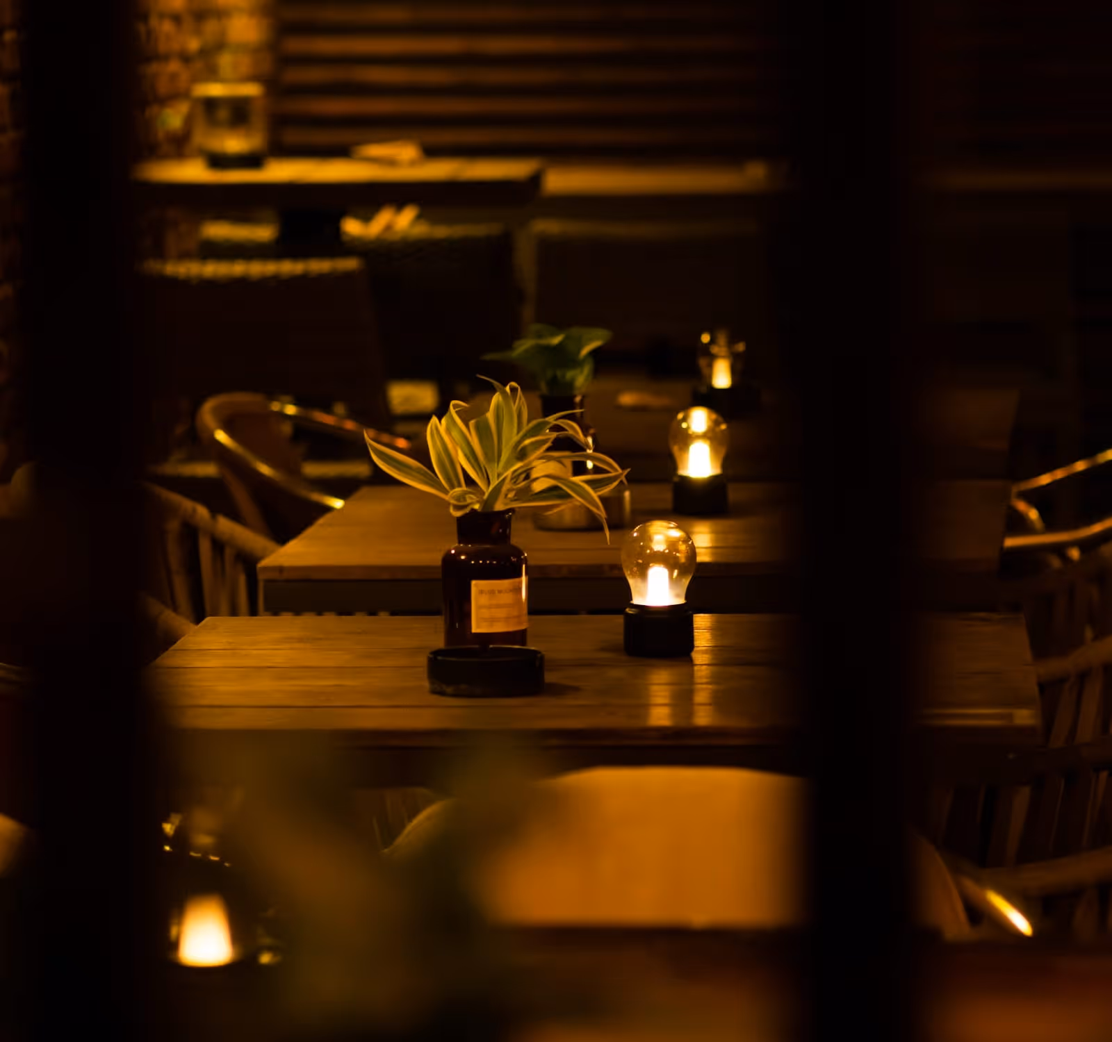 The dining room after dark, wood tables set with small brass lamps against the bare brick wall