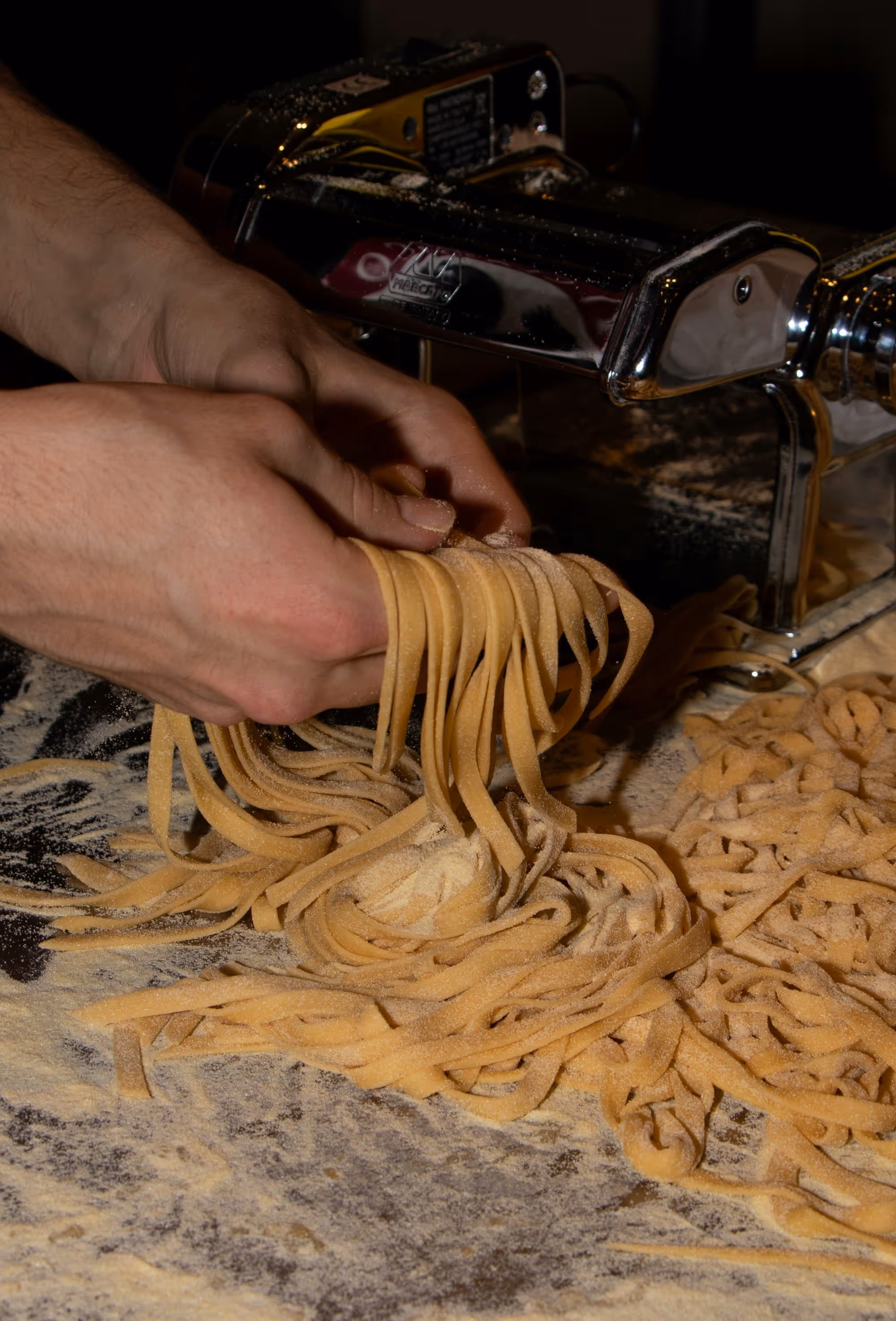 Hands lifting fresh-cut pasta away from the roller onto a semolina-dusted board